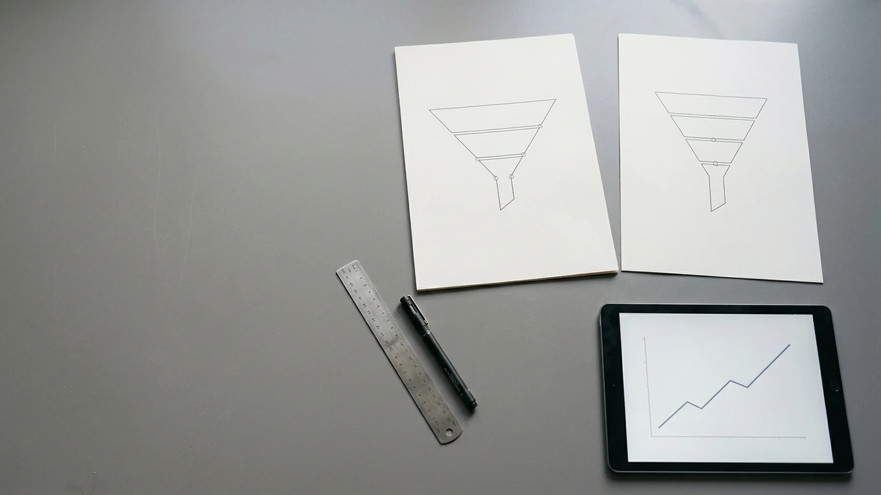 Desk with two printed funnel charts side by side, a ruler and a pen, and a tablet showing a line graph in cool daylight
